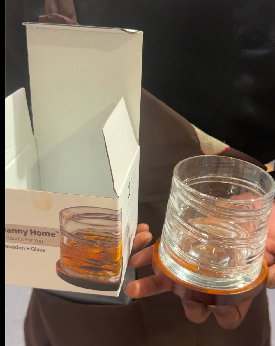 Clear round whiskey glass with a wooden base, held by hand beside its Danny Home packaging box.